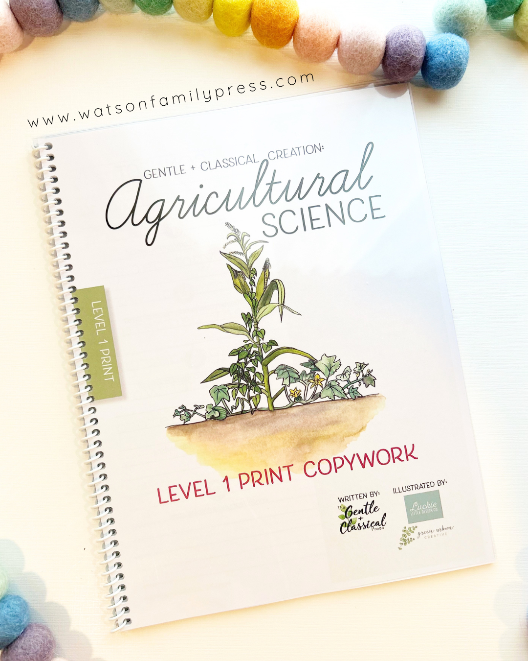 A spiral-bound copywork book titled 'Agricultural Science' from Watson Family Press, featuring a watercolor illustration of a plant in a pot on the cover, photographed on a light background with colorful pom-poms along the top edge.