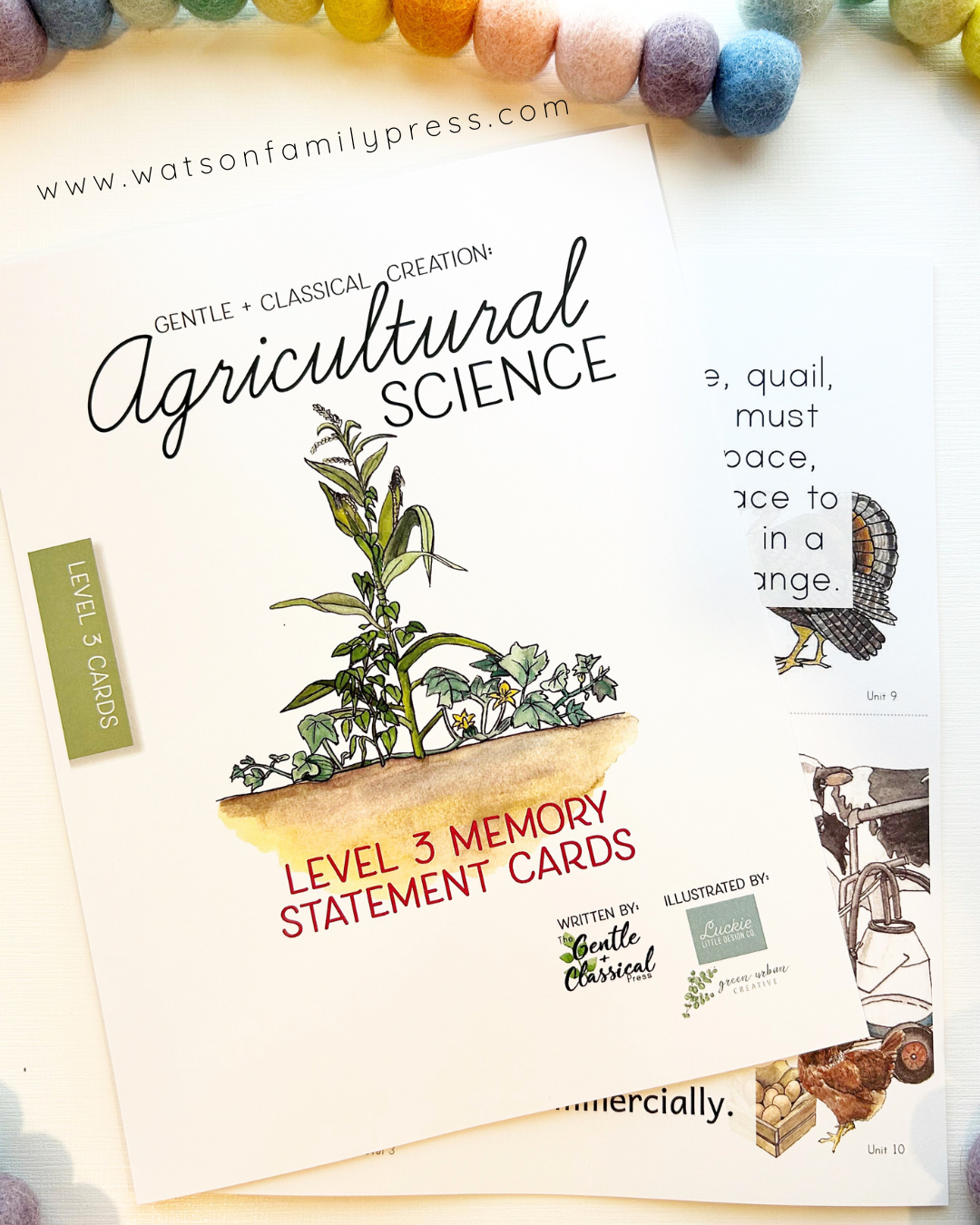 A set of printed memory statement cards titled 'Agricultural Science' from Watson Family Press, arranged on a light surface with colorful felt pom-poms along the top edge. The cards feature illustrated botanical and animal drawings, text, and decorative elements.