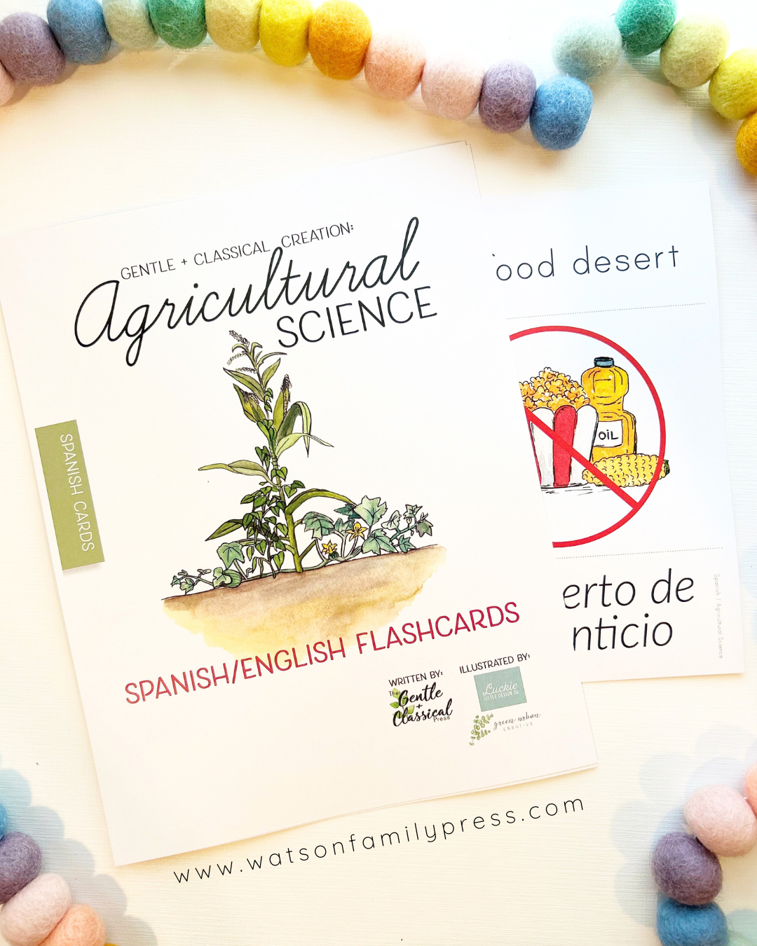 Spanish/English flashcards for Agricultural Science displayed on a white surface, surrounded by pastel pom-poms. The main card features a watercolor illustration of a plant and the title 'Agricultural Science'.