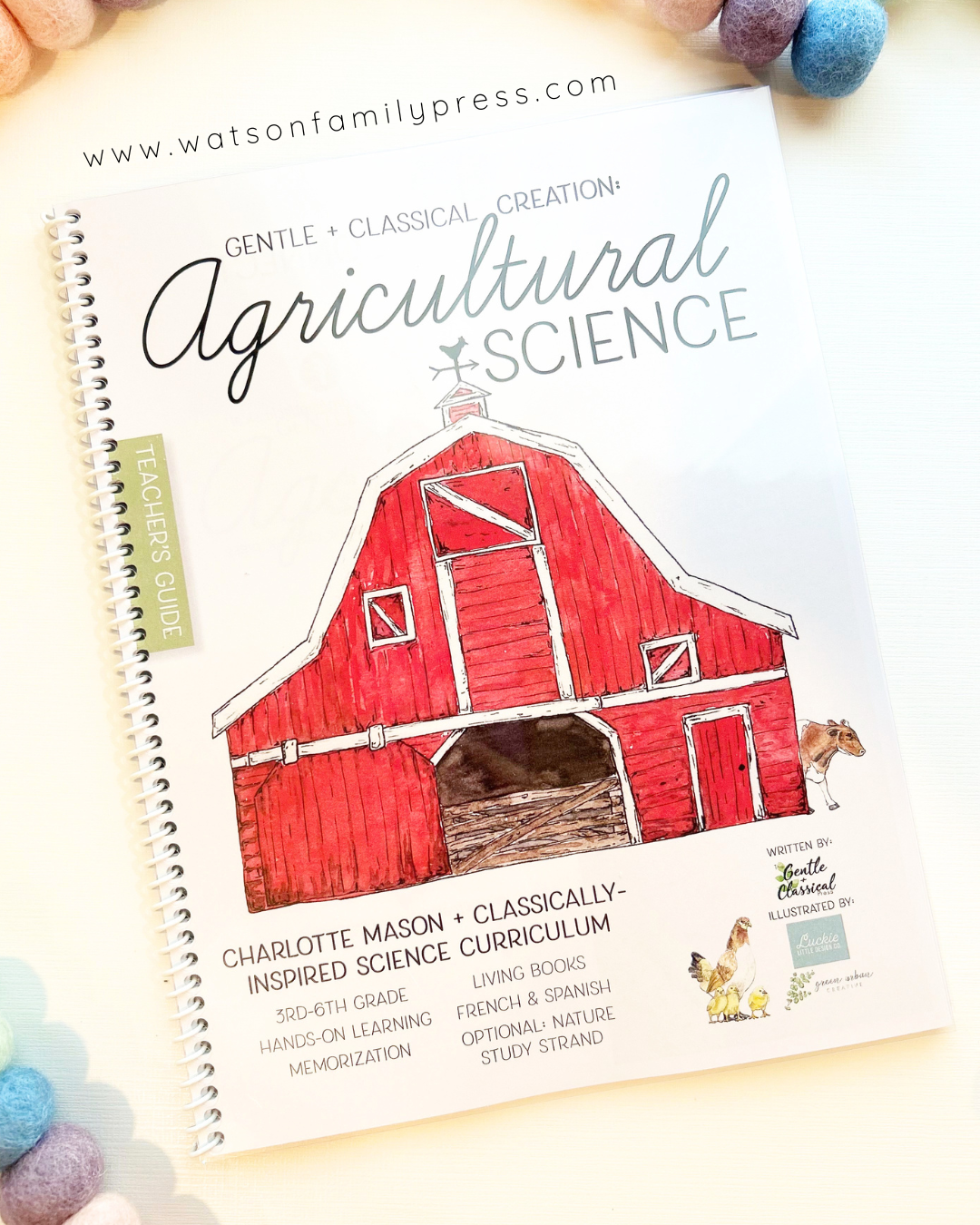 A spiral-bound book titled 'Agricultural Science' from Watson Family Press, featuring a red barn illustration on the cover, photographed on a light background with colorful pom-pom accents around the edges.