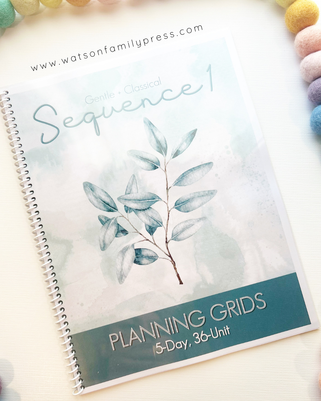 Spiral bound book entitled 'Gentle + Classical Sequence 1 Planning Grids' with a branch illustration on the front cover on a light surface with colorful pom-pom beads around it