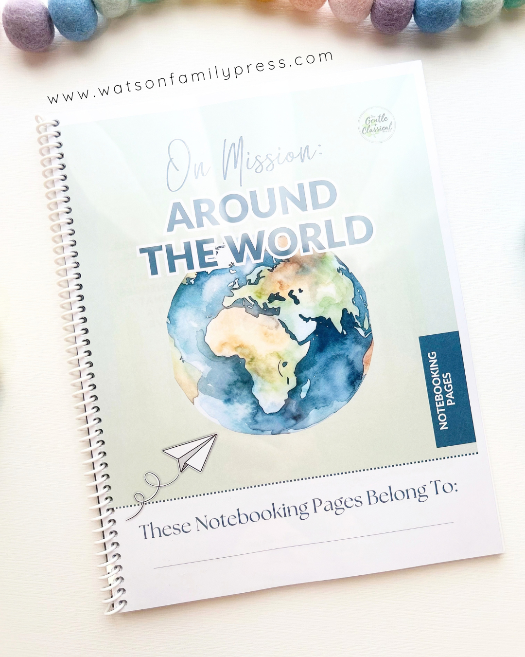 A spiral-bound educational curriculum booklet titled 'On Mission Around the World' with an earth illustration on the cover, photographed on a light surface with colorful felt pom-poms around it.