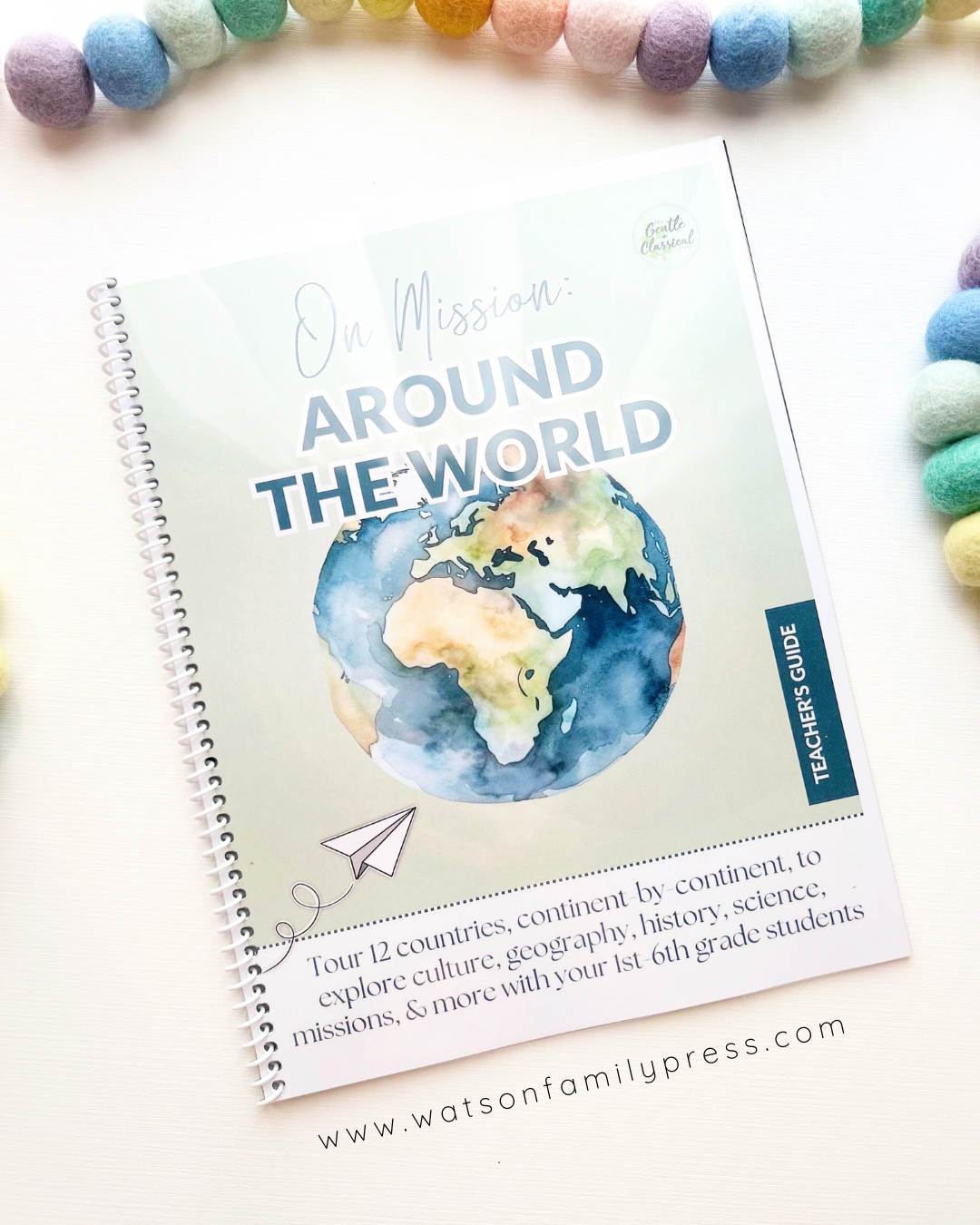 A spiral-bound educational curriculum booklet titled 'On Mission Around the World' with an earth illustration on the cover, photographed on a light surface with colorful felt pom-poms around it.