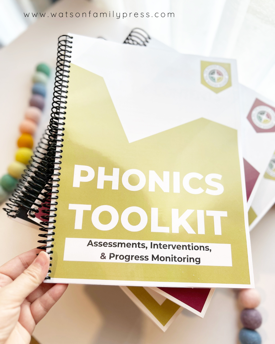 A spiral-bound educational curriculum booklets stacked, titled 'Phonics Toolkit' with a yellow front cover, photographed on a light surface with colorful felt pom-poms around it.