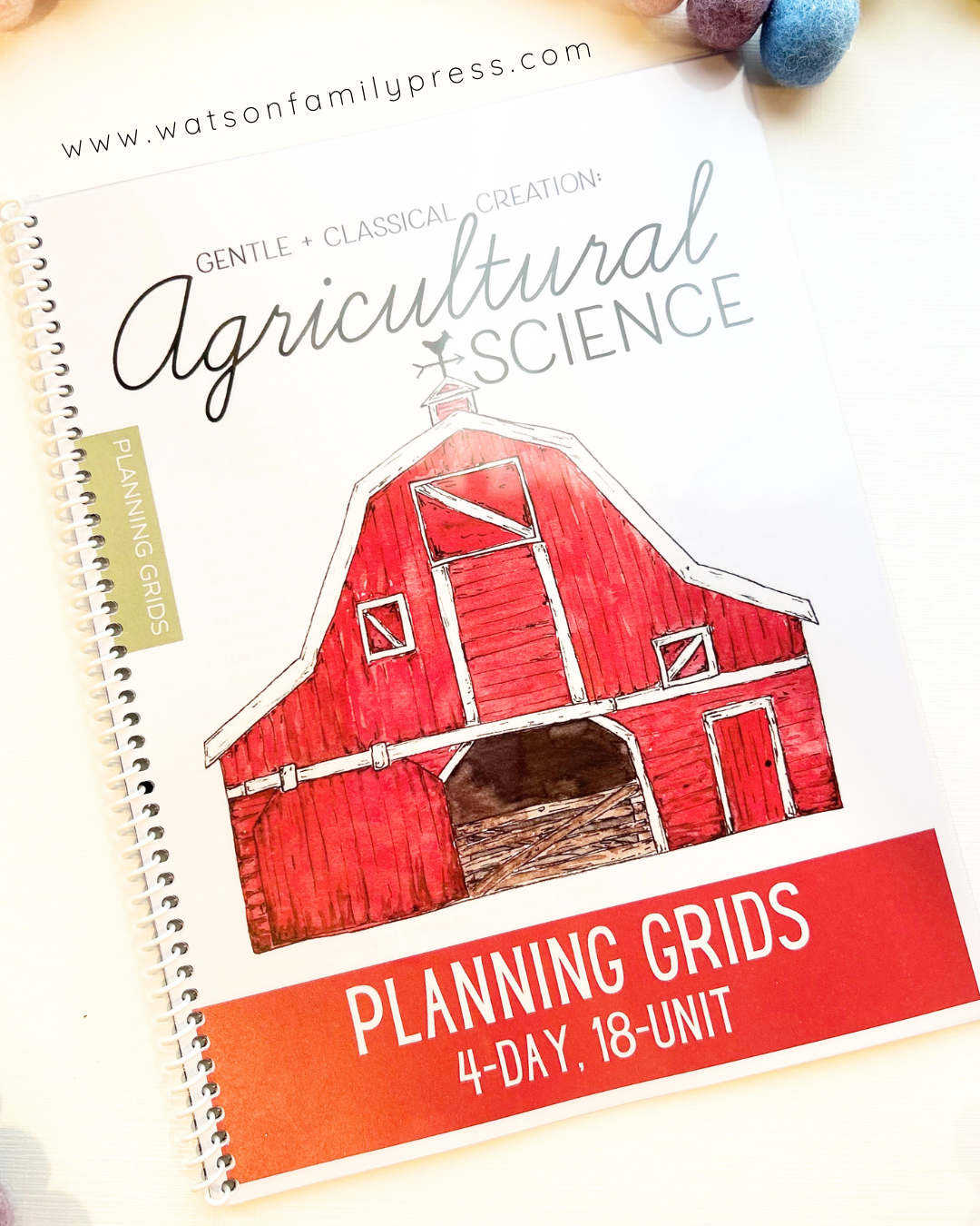 A spiral-bound planner pad titled 'Agricultural Science Planning Grids' with a red barn illustration on the cover, photographed on a light surface with small colorful decorative objects at the top edge.