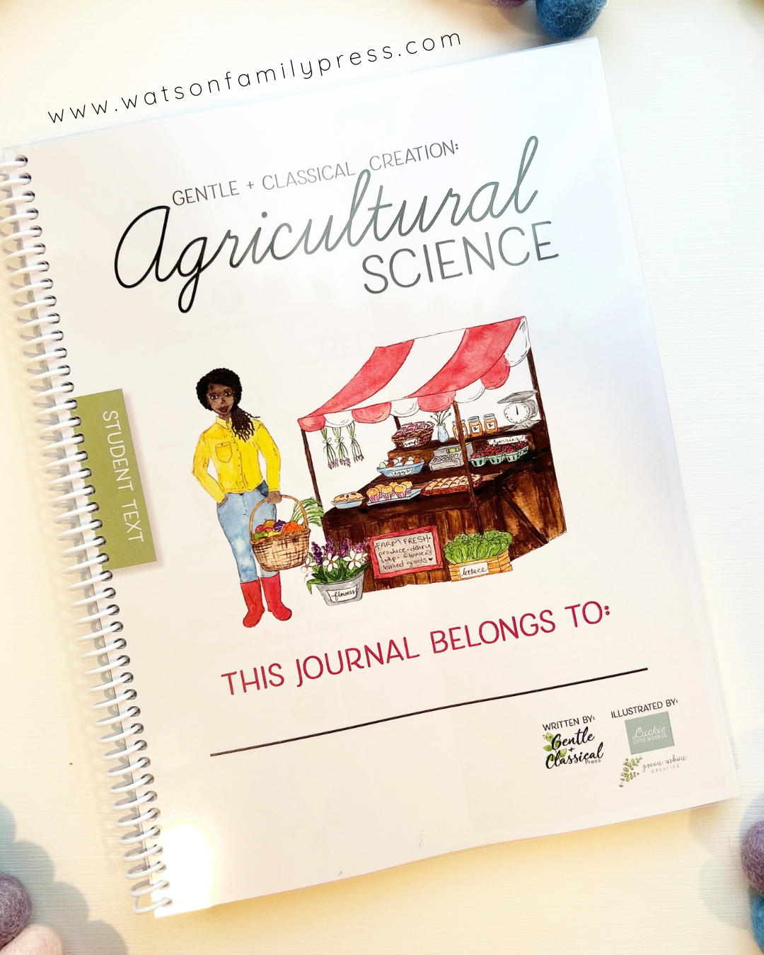 A spiral-bound journal titled 'Agricultural Science' from Watson Family Press, featuring a vintage-style illustration of a farmer at a market stall. The journal is photographed on a light surface with colorful pom-pom props in the corners.