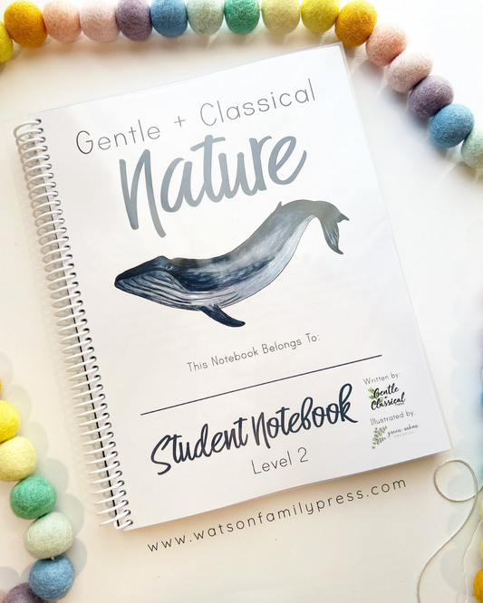 A spiral-bound notebook titled 'Gentle + Classical Nature' with a whale illustration on the cover, styled on a light surface with colorful pom-pom beads around it. Visible text includes 'Student Notebook Level 2' and 'www.