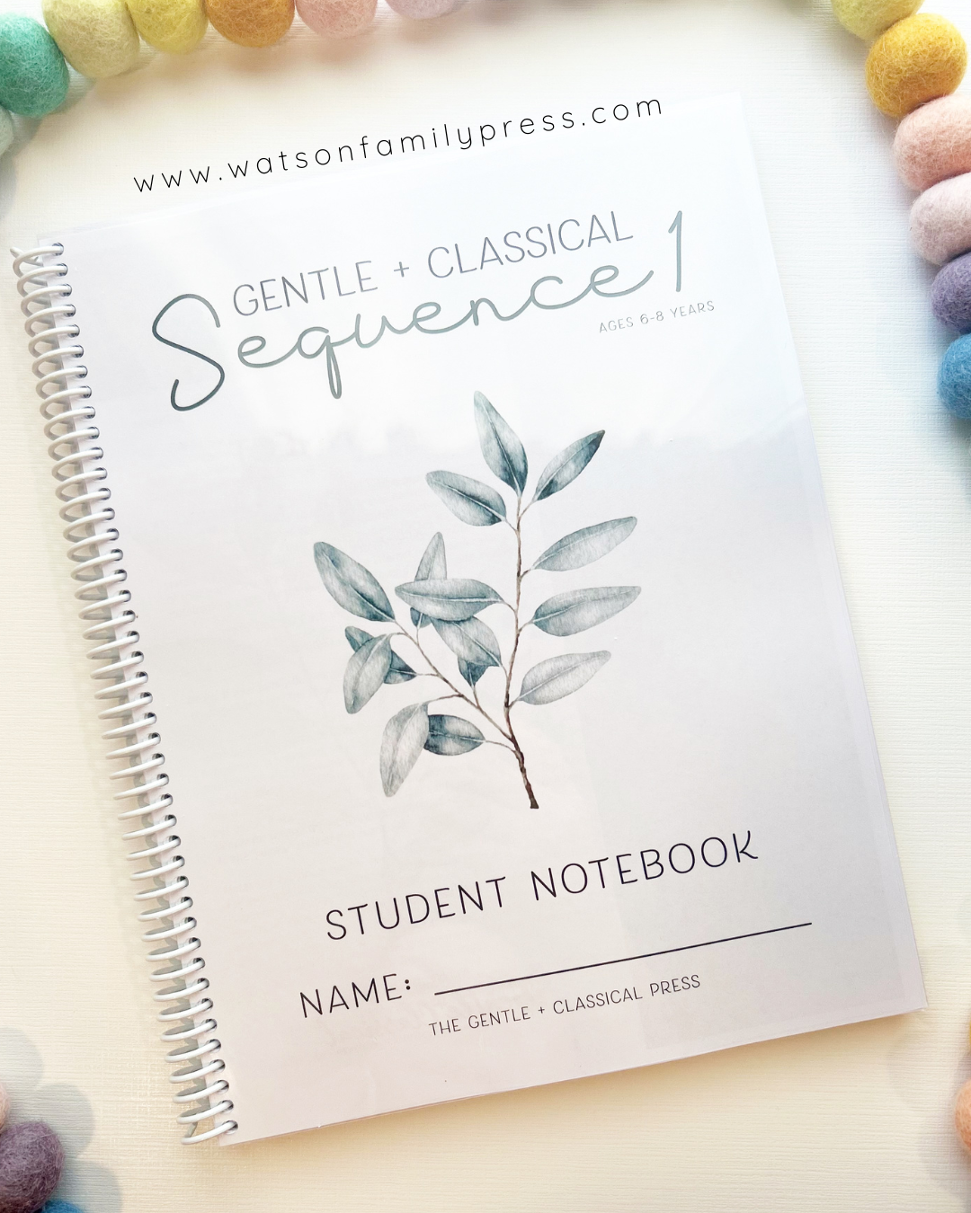 A spiral-bound student notebook titled 'Gentle + Classical Sequence 1' with a watercolor leaf illustration on the cover, photographed from above on a light surface with colorful pom-pom beads around the edges. 