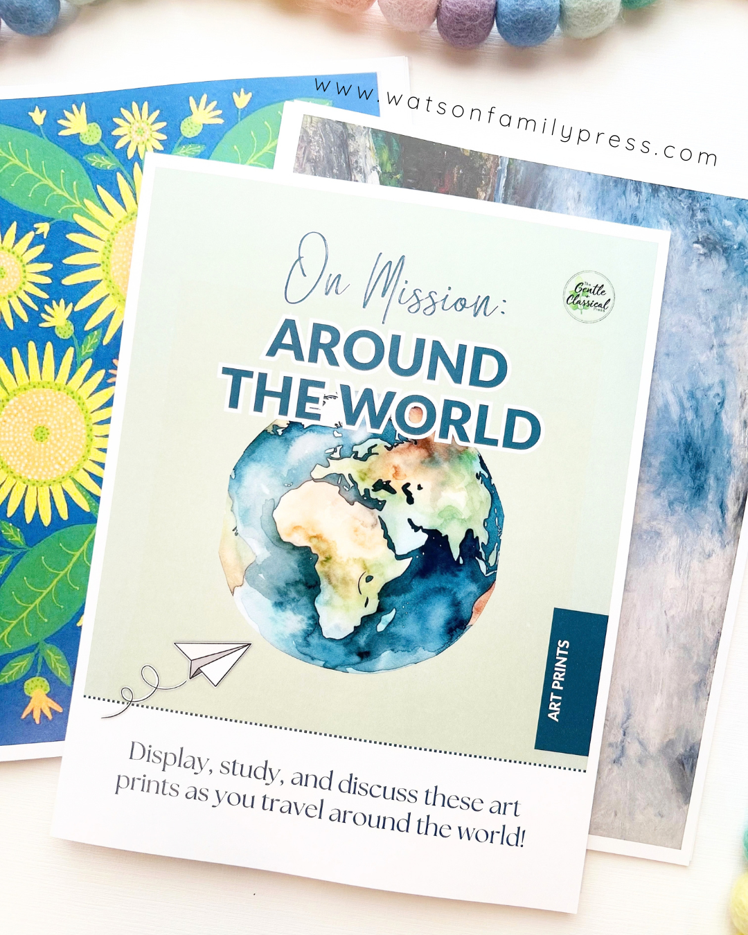 A loose leaf educational curriculum booklet titled 'On Mission: Around the World' with an earth illustration on the cover, photographed on a light surface with colorful felt pom-poms around it.