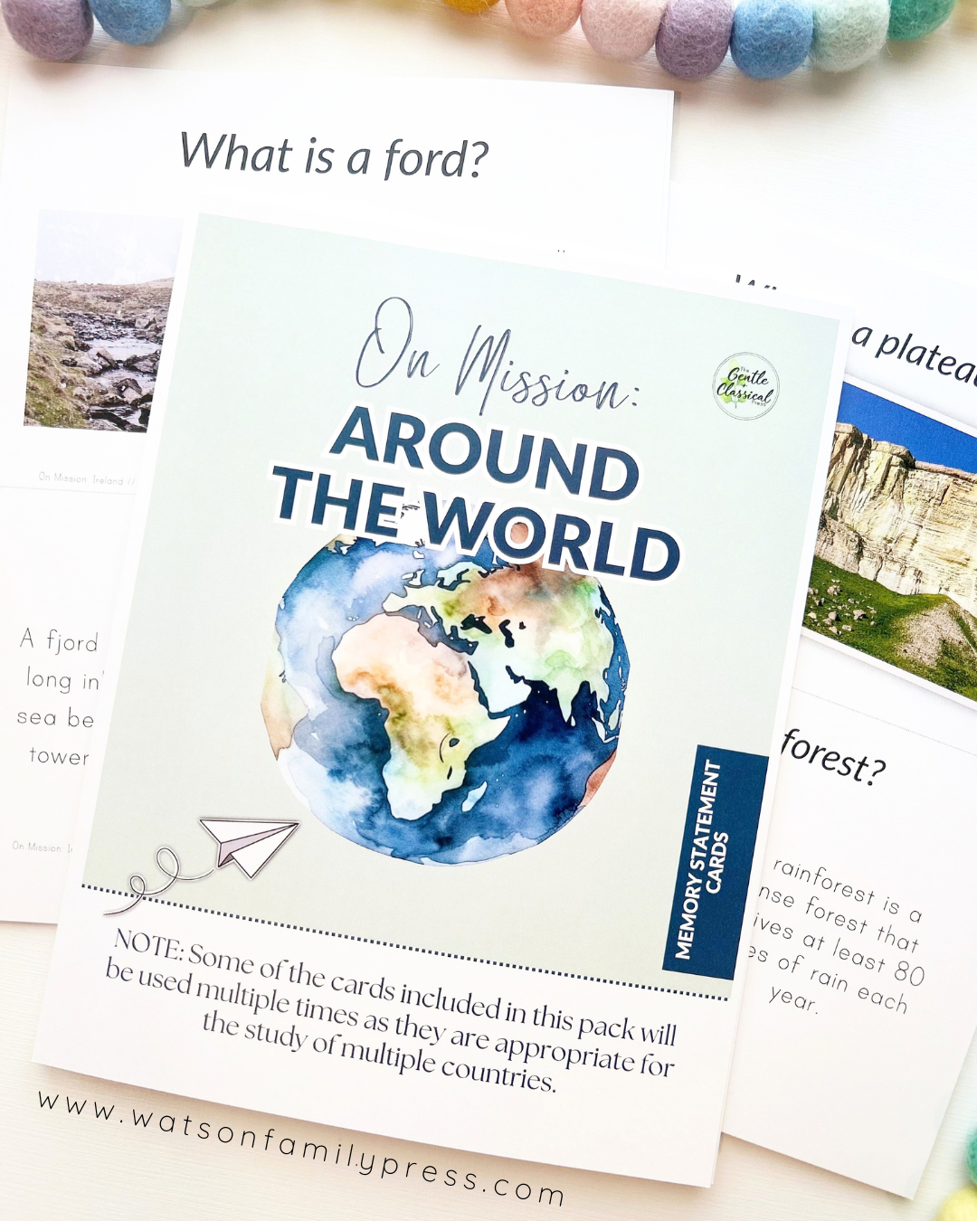 A loose leaf educational curriculum titled 'On Mission: Around the World' with an earth illustration on the cover, photographed on a light surface with colorful felt pom-poms around it.