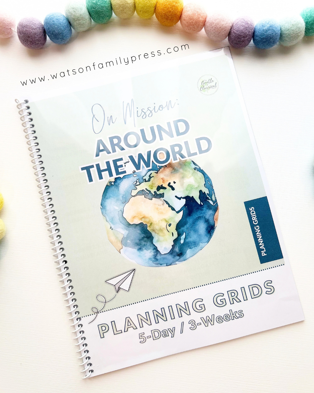 A spiral-bound educational curriculum booklet titled 'On Mission Around the World' with an earth illustration on the cover, photographed on a light surface with colorful felt pom-poms around it.